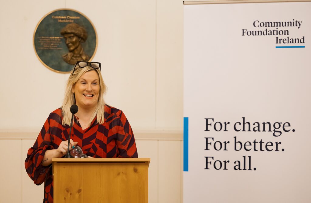 A woman standing at a podium, smiling with a bronze plaque behind her on a wall. To the right of the photo is a pull up banner of Community Foundation Ireland. This is Lisa-Nicole Dunne, Chief Executive of Mantra Strategy.