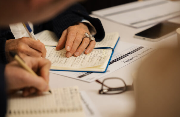 A photo of hands note-taking at a desk. You can see three hands, two of which are holding pens writing into note pads. This is from a conference environment.