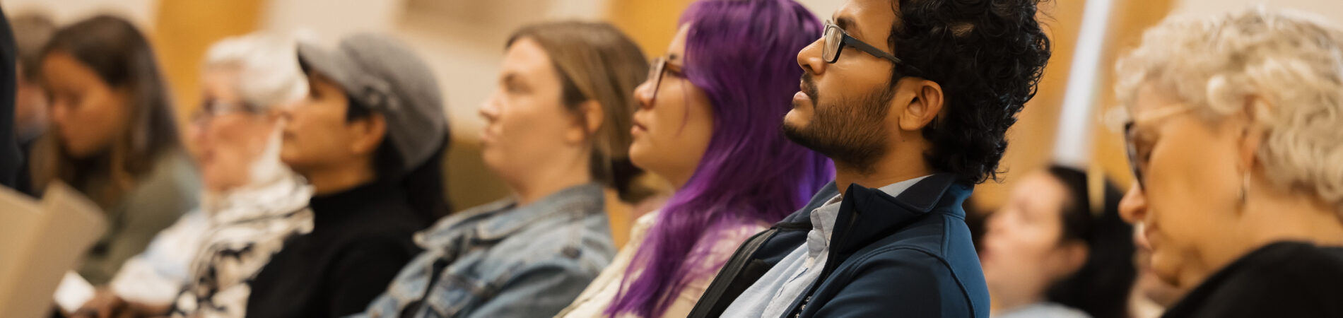 A row of people sitting, shot from the side they are in an audience at an event and facing towards the left of the picture.