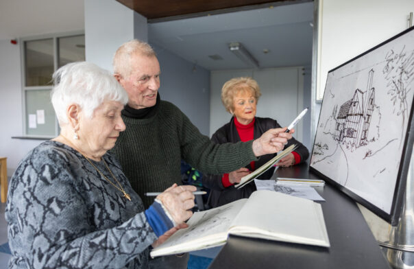 Three older people at a drawing class, two women with a man at the centre are looking and amending a drawing. The scenic drawing is on the right of the photo. They are looking at it and amending it.