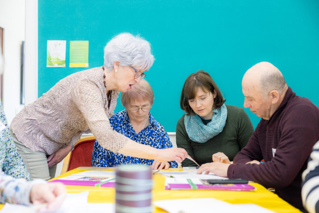 Four adults - one standing on the left, and three seated at a table. They are working together. The lady standing is pointing at notes on the table.
