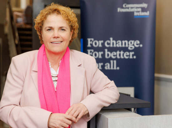 A woman dressed in a pink jacket and darker pink scarf, looking at the camera. Denise Charlton is standing in front of a Community Foundation Ireland banner which says 'For change. For better. For all.'