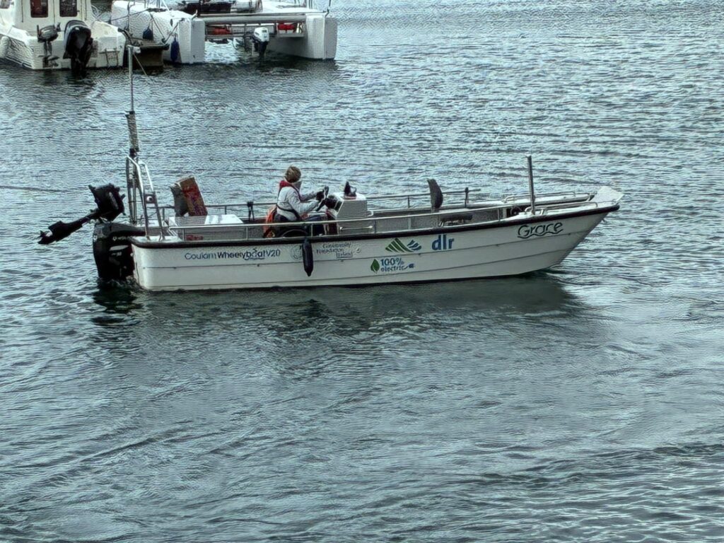 Accessible boat Grace, afloat in Dun Laoghaire Harbour. She is an open boat with a white hull - on the side are the logos of the partners. The boat is powered by an out-board engine in the rear which is visible.