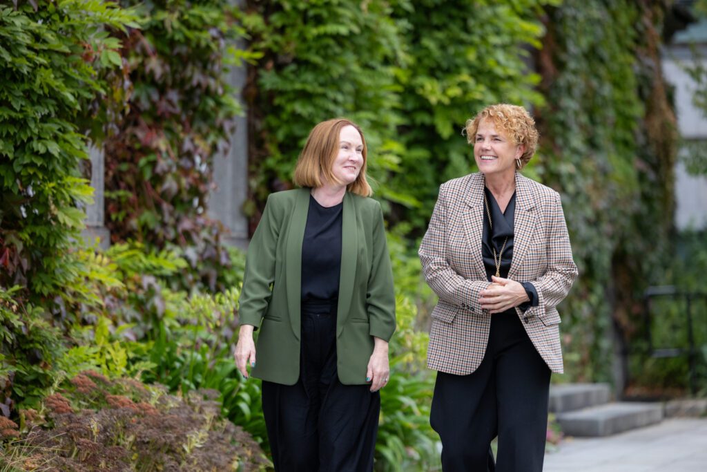 Walking past a building with green ivy clad walls are two individuals, Moninne Griffith and Denise Charlton. Both are smiling as Moninne prepares to take up new role as Director of Impact, Grants and Donor Care at Community Foundation Ireland.