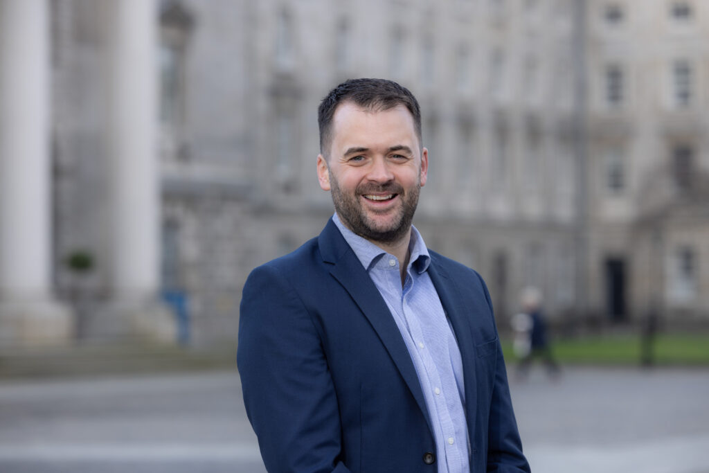 A man facing the camera in an open necked shirt and business jacket. John Sansome is in the courtyard of Trinity College Dublin - with its pillared buildings blurred as a backdrop.