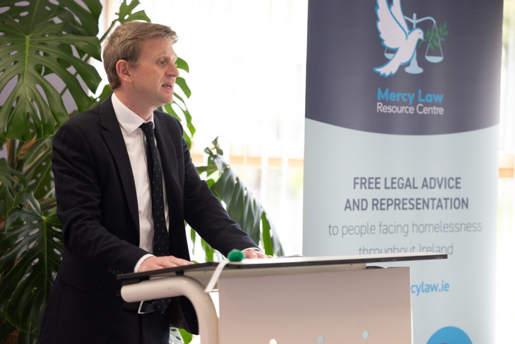 A man speaking at a podium with the backdrop saying the Mercy Law Resource Centre. Paul Dornan of the Centre is speaking at the launch of the research.