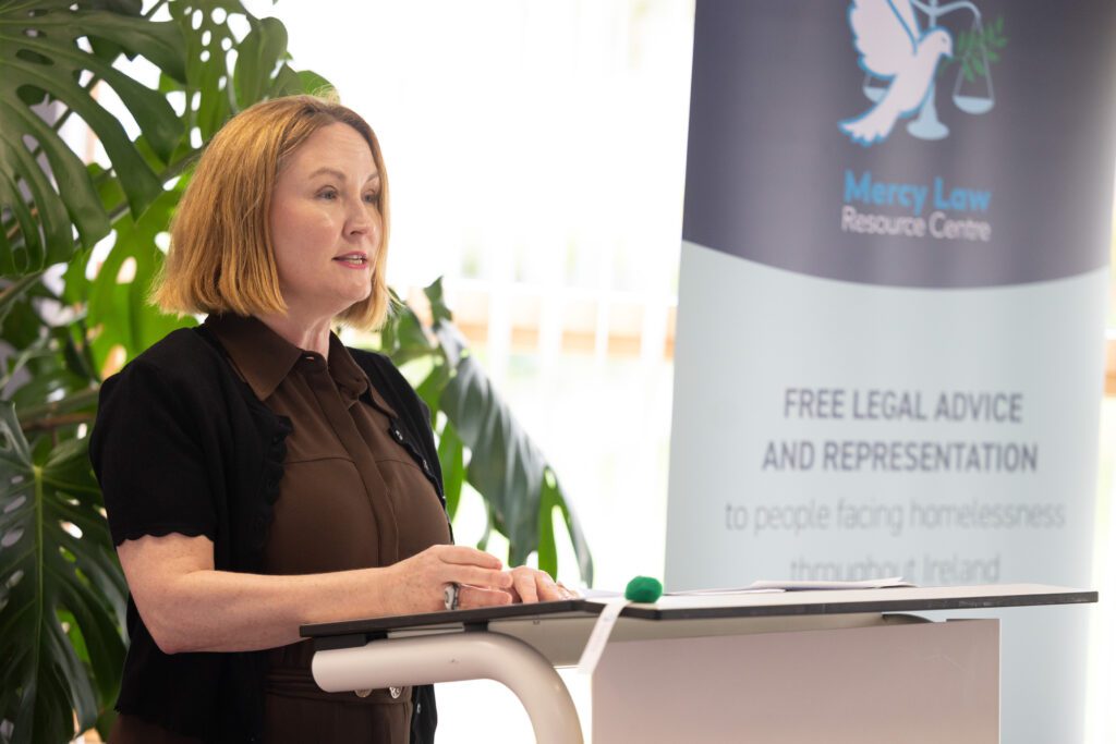 A picture of a woman at a podium, with a back drop of a banner with the logo of the Mercy Law Resource Centre. Moninne Griffith of Community Foundation Ireland is speaking at the launch of pioneering research.