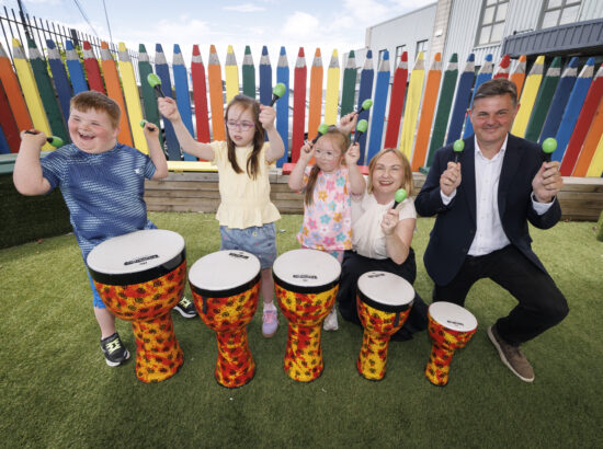 Against a backdrop of the posts of a multi coloured fence, three children from Down Syndrome Centre are playing with musical instruments including Bongo drums, on their immediate left is Moninne of Community Foundation Ireland and Kevin from RTÉ