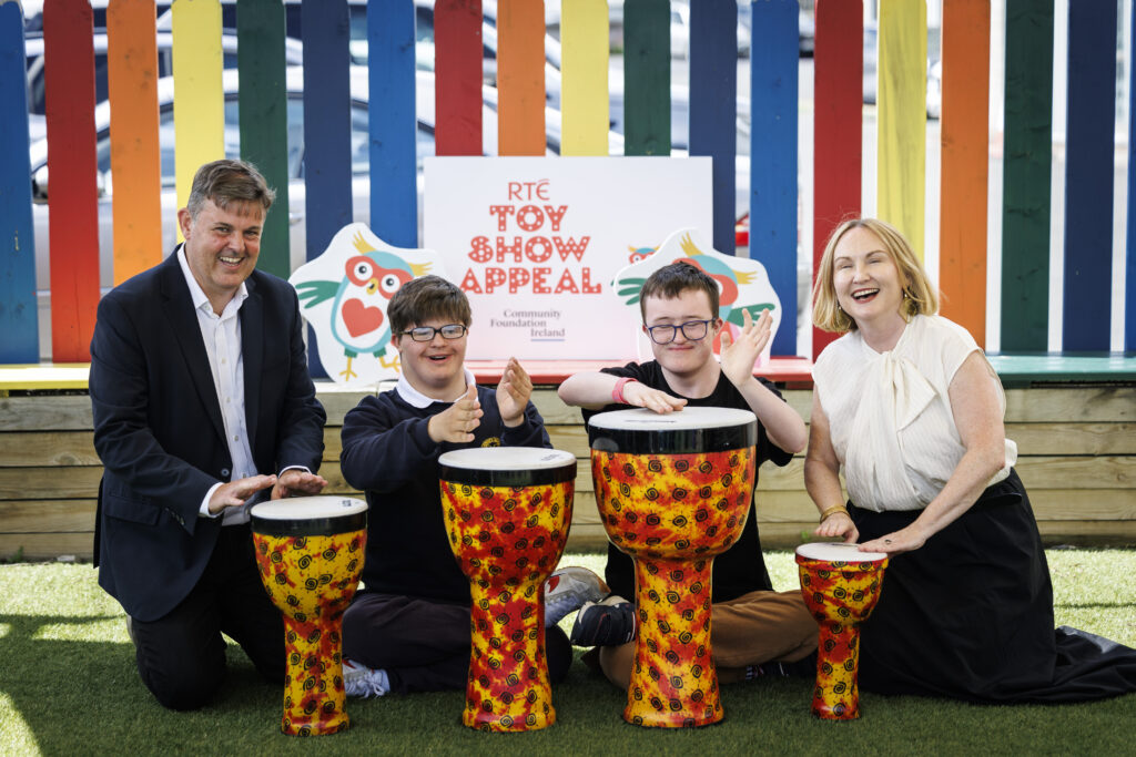 Against a backdrop of a multi coloured fence, four people are banging drums. Two children from Down Syndrome Centre are in the centre, with Kevin Bakhurst of RTÉ on the left and Moninne Griffith of Community Foundation Ireland on the left.