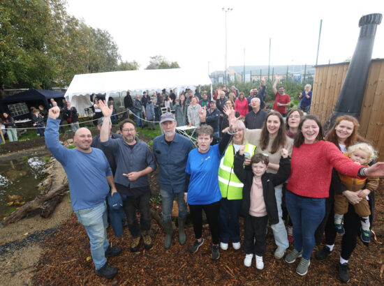 A large group of people in a community garden, the are in outdoor clothing, smiling and waving their arms at the camera.