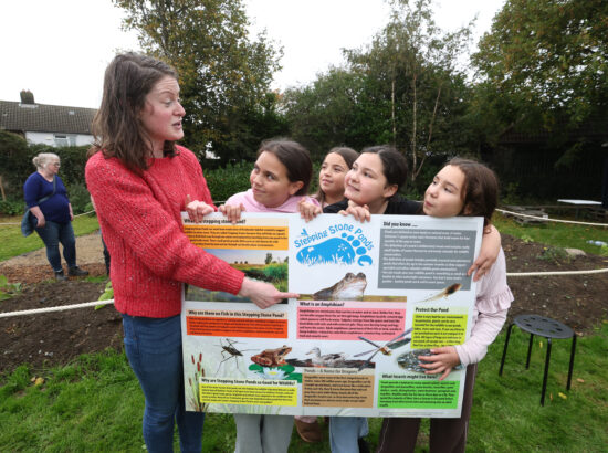 With the background of a garden, four girls are holding up a large board which is a guide to Stepping Stone Ponds. A woman to the left of the photo is pointing and explaining the contents.