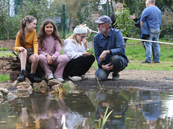 Three girls sitting on low rocks next to a pond, with reflections on the water. They are being told about nature by a man in outdoor clothes and a cap. This is one of 300 communities across Ireland with a local biodiversity action plan as a result of a partnership with Community Foundation Ireland.