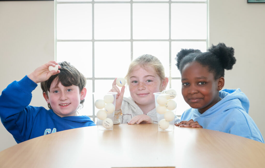 Three children playing with a glass and ping-pong balls on a table.