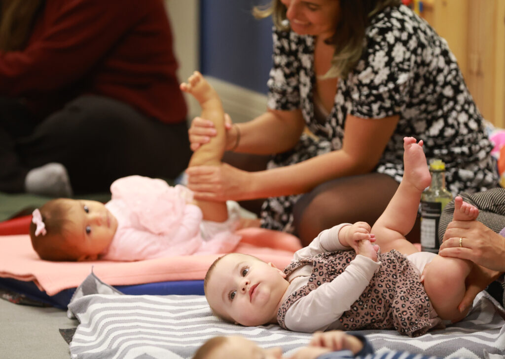 Two babies lying on mats on the floor. They are looking towards the camera. Adults are massaging their legs which are raised.