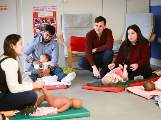 This is a group of parents - there are three babies on mats in a community room setting. At their feet are three crouching adults - parents or guardians. An instructor on the left of the picture is using a model of a baby to show how to massage their legs to promote development.