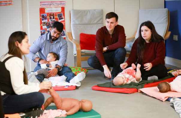 Four adults with three babies lying on floor mats. The adults are crouching down and massaging the babies. On the left is an instructor with a model giving instructions.