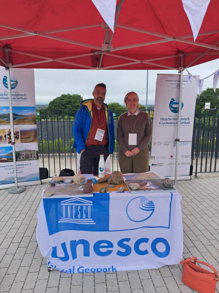A man and a woman at a stall, draped over a table and facing forward is a cloth with the UNESCO logo, AIlbhe from Community Foundation Ireland is meeting Ben Thébaudeau, Joyce Country and Western Lakes UNESCO Geopark.