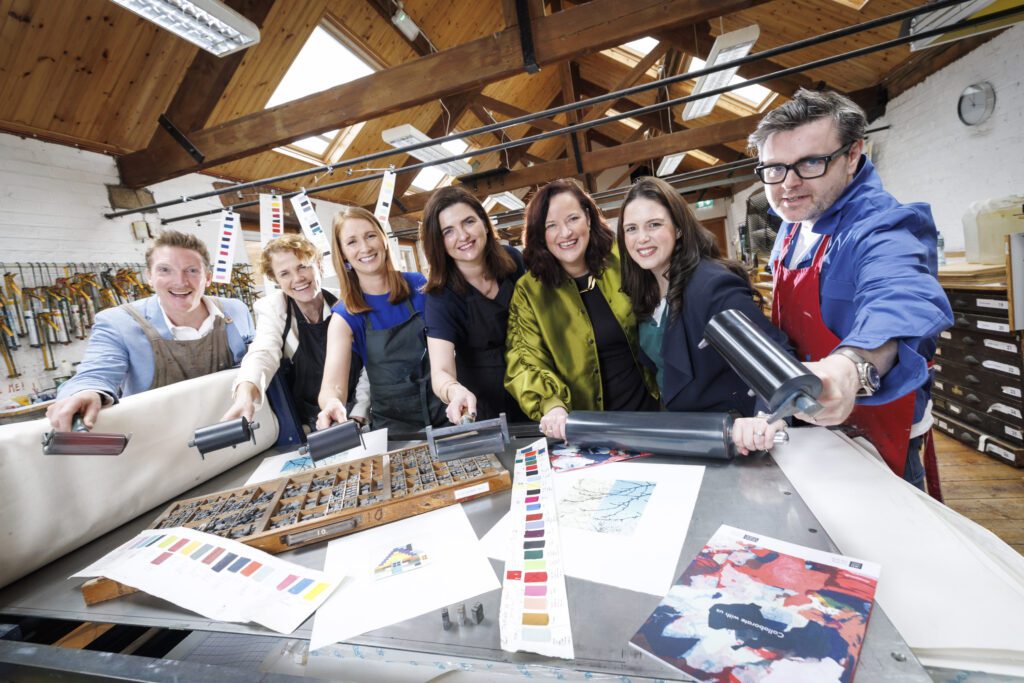 A group of people, partners of Business to Arts in a graphic studio. They are overlooking a bench filled with graphic design material.