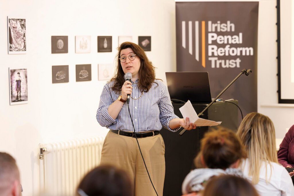 A woman speaking to a meeting, she is holding a microphone and has a piece of paper in her left hand. In the background is a pull up banner of the Irish Penal Reform Trust.