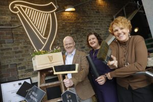 ere’s the suggested alt text for the image: "Three people standing in a well-lit room with a neon sign reading 'We Dream in Beer' in the background. One person holds a microphone and a graduation cap. A wooden crate filled with flowers and branded with the Guinness logo is in front of them, along with a can of Guinness on the table."