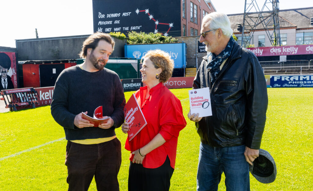 On the pitch at Dalymount Park are three people. Sean McCabe of Bohs is on the left, In the middle is Denise Charlton of Community Foundation Ireland she is holding a copy of the new Strategy for the Bohemian Cooperatives with a red cover on the right is Ted Howard of Democracy Collaborative. In the far background is a mural of the Plough and Stars painted on a gable of a building - a traditional Irish symbol of workers rights.