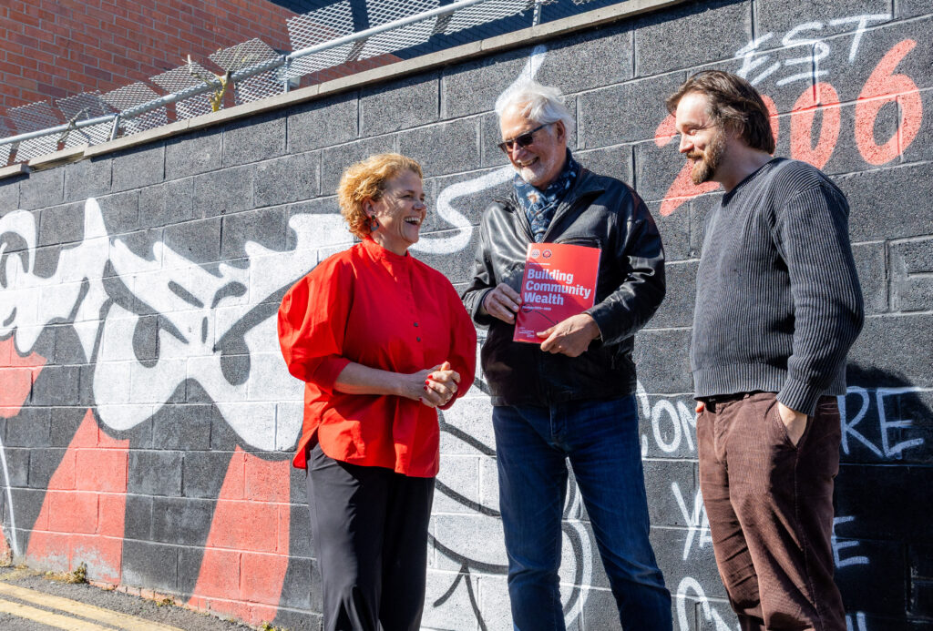 Three people against a backdrop of a wall with a black, red and white mural. They are in discussion. Denise Charlton on the left is in discussion with Ted Howard of the Democracy Collective and Sean McCabe of Bohs. Ted in the middle is holding a copy of the strategy for Bohemians Cooperatives. 