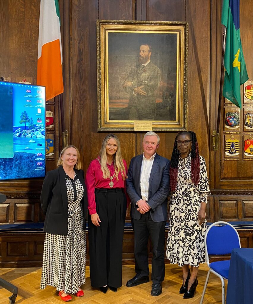 A group of four people, three woman and a man second from left. They are the speakers for the launch of the documentary 'Echoes of 2004'. They are standing with a backdrop of a portrait painting and wooden panelled wall. This is the Oak Room of Dublin's Mansion House. On the left side of the portrait on the Irish tricolour and the flag of Dublin.