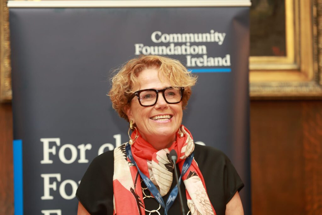 Denise Charlton is speaking at a podium. The photo is head and shoulders she is wearing a black top and a red scarf, behind her is Community Foundation Ireland branding including a logo on a pop up banner.