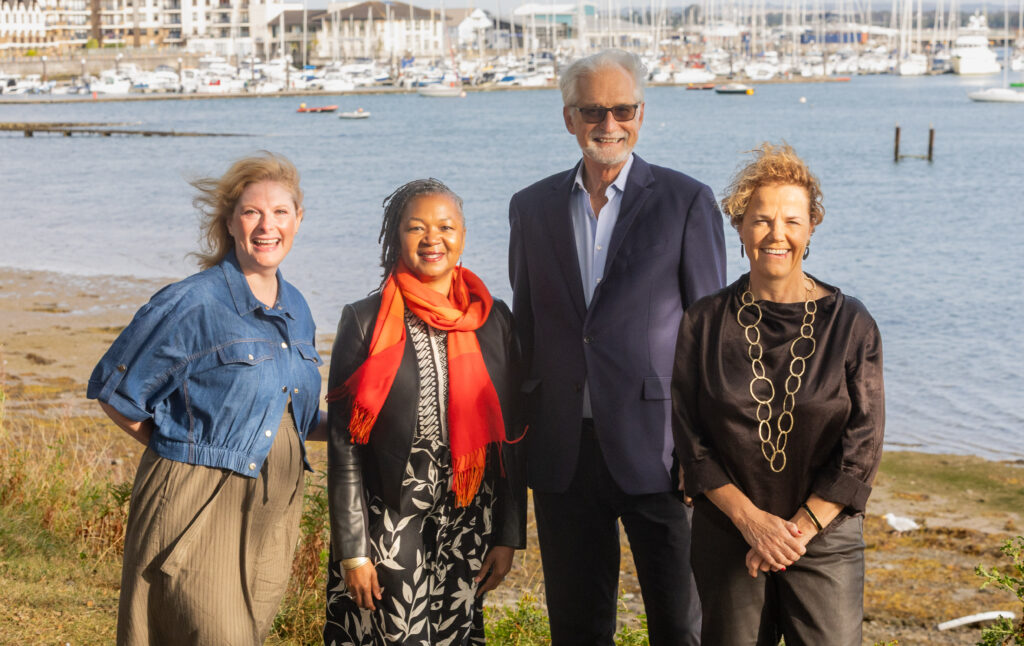 Four delegates of the Global Summit of Community Foundation Leaders in Malahide Dublin, the backdrop is the sea and a marina of boats. On the left in a blue jacket is Sharon Avery of Toronto Foundation, Tonia Wellons of Greater Washington Foundation, Ted Howard of Democracy Collaborative and Denise Charlton of Community Foundation Ireland.