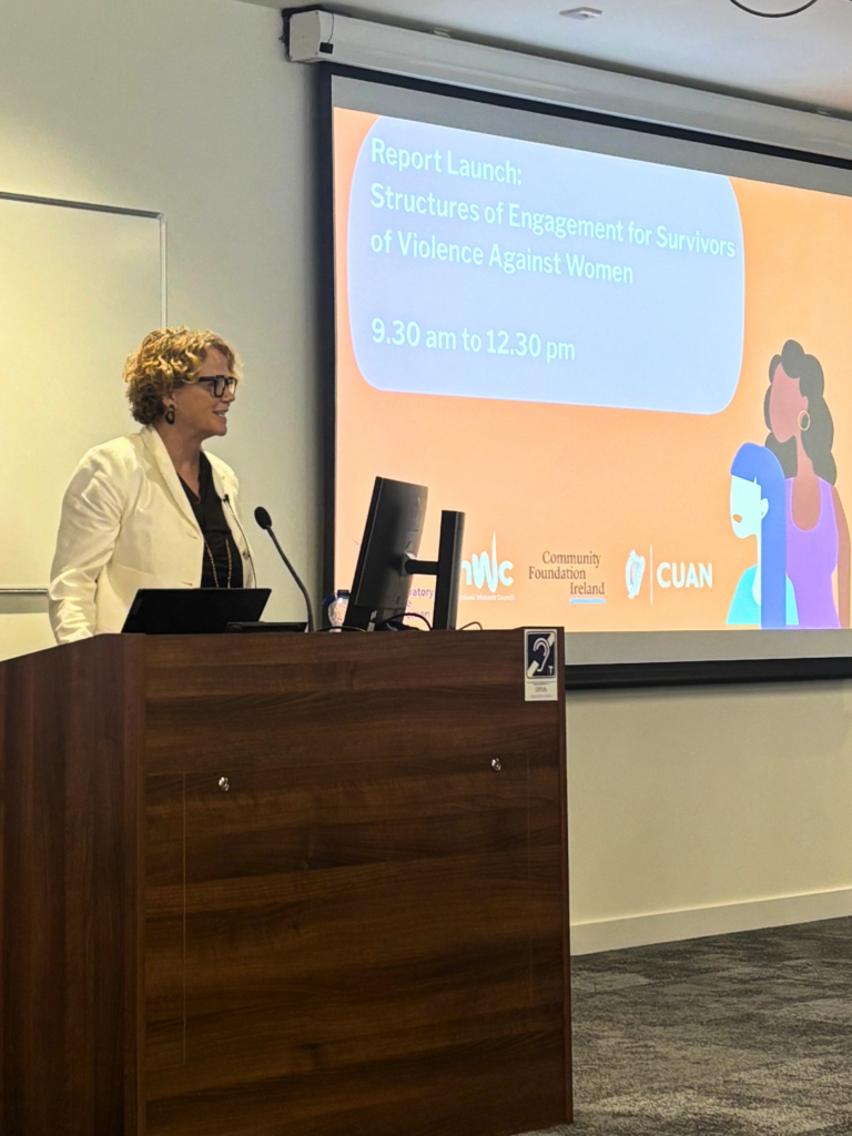 A woman in a white business jacket at a podium. To her right is a presentation scheme. Denise Charlton is speaking at the launch of the research.