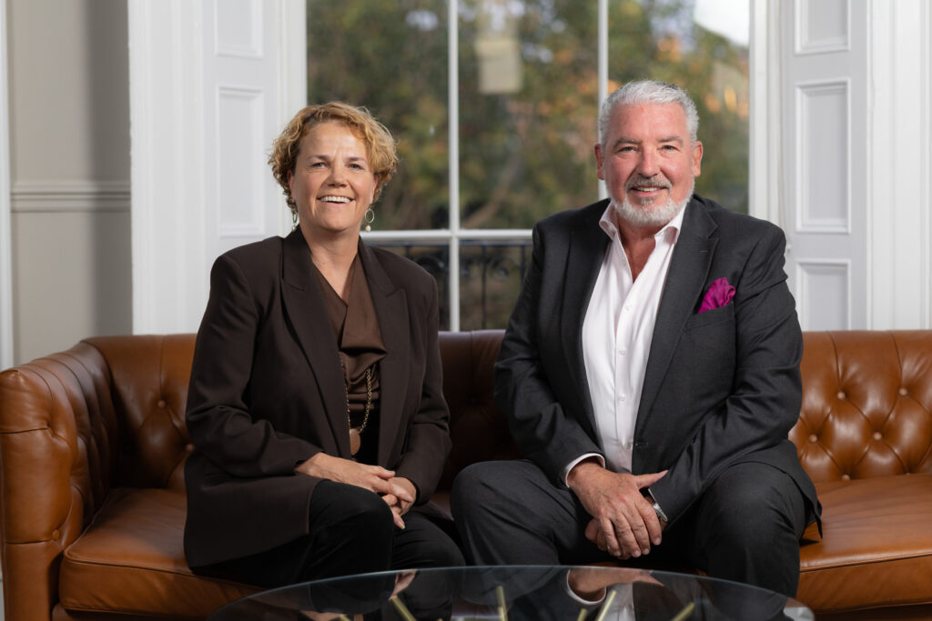 A man and a woman sitting on a leather sofa. Denise Charlton, Chief Executive of Community Foundation Ireland is welcoming new Chair John Cunningham. The background is a window where trees in autumn foliage are visible. Both are looking at camera and smiling.