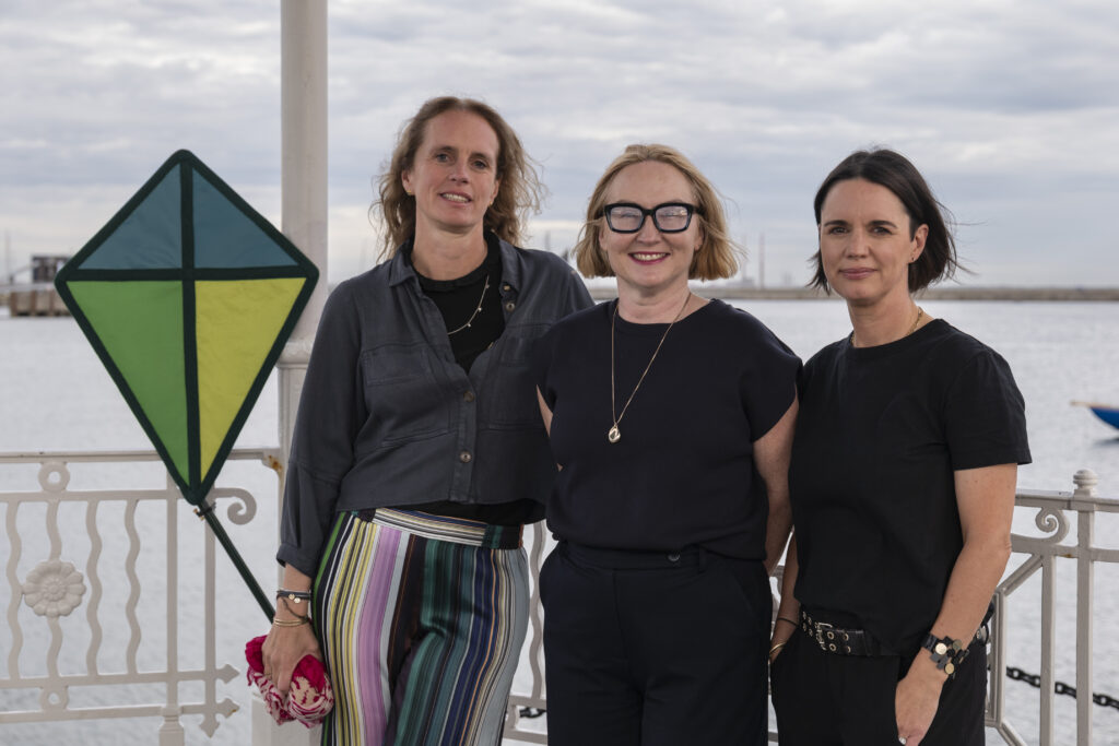 Three woman facing the camera - representatives of Green Kite and Community Foundation Ireland they are smiling - to their immediate left is a kite. In the background is Dun Laoghaire Harbour, Dublin.
