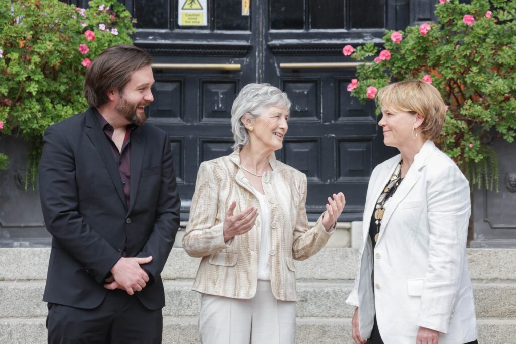 Three people in conversation on steps leading to the entrance to a building. On the left in dark colours is Sean McCabe of Bohs FC. In the centre President of Ireland, Catherine Conolly who has her hands raised and is turned to her left talking to Denise Charlton of Community Foundation Ireland. The President is arriving for the launch of Bohemian Cooperatives.