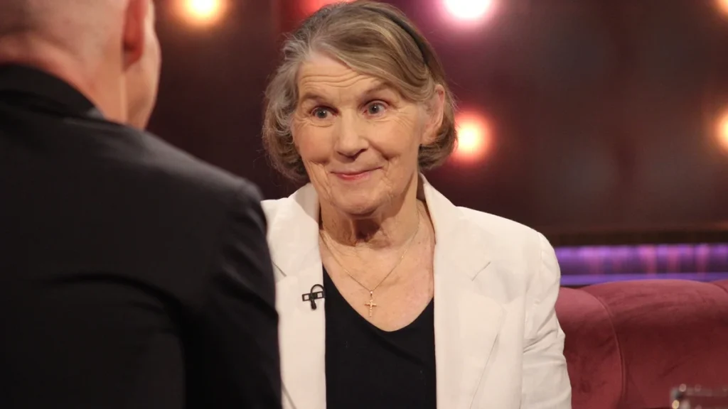 Picture of Sr Stan sitting dressed in white jacket in what is a studio situation. The image also captures the back of a male interviewer.