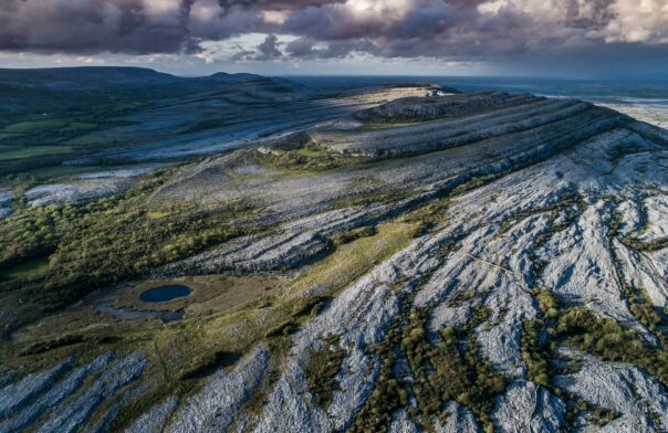 Aerial photo of the Burren, Co Clare. A limestone landscape with plant life emerging from crevices is set against a backdrop of distant mountains - under a cloudy sky.