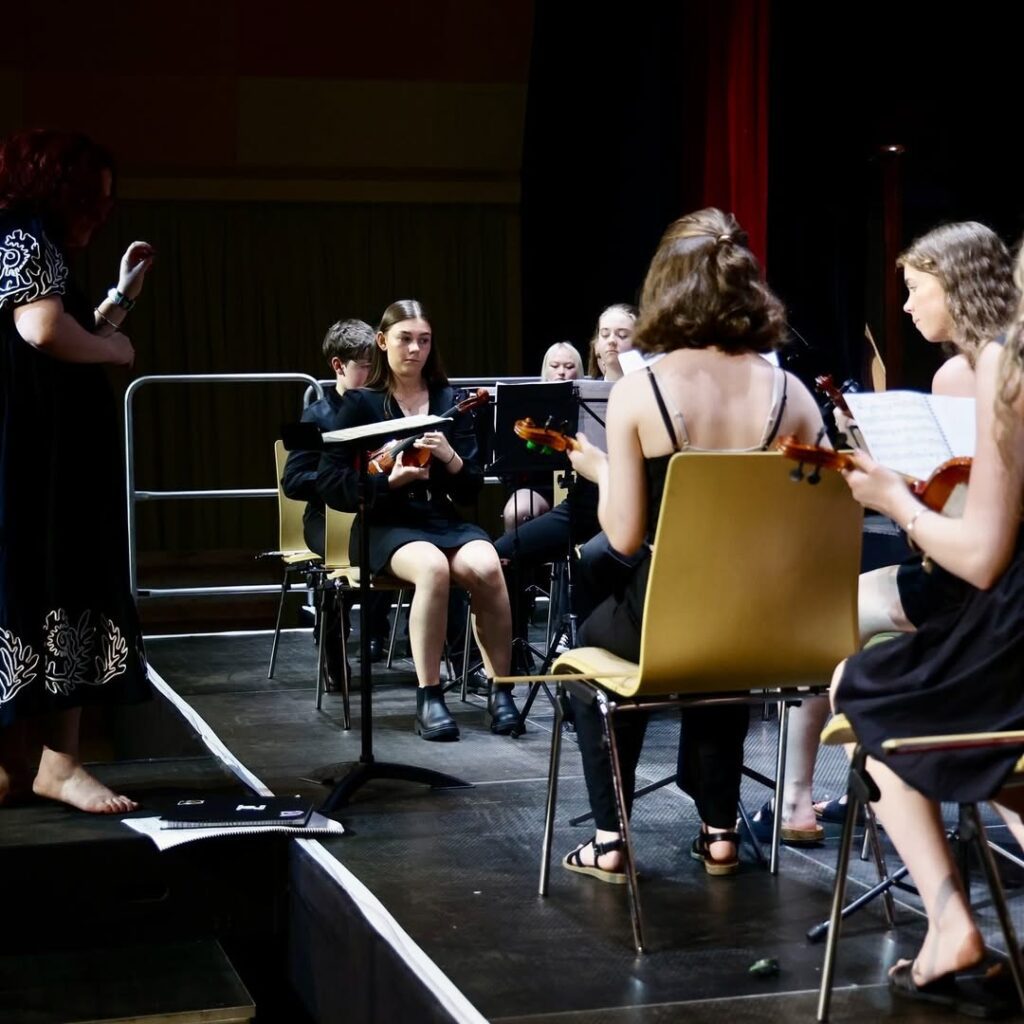 Three young musicians on stage. They seem to be part of an orchestra. Two have their backs to the camera while a third is playing a violin., They are set against a black backdrop. These are members of the Irish Chamber Orchestra.