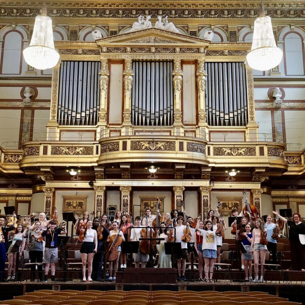 An Orchestra of 30 players in a grand opera house style setting. They are standing on a stage holding instruments against a backdrop of organ pipes and two chandeliers. These are young members of the Irish Chamber Orchestra on a trip of a lifetime to Vienna