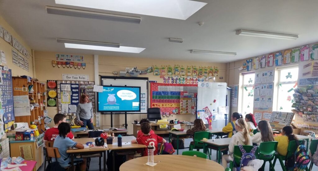 A school room environment taken from the back of a class. The backs of young students can be seen, they are sitting facing a screen. A teacher is explaining about whale and dolphin strandings. This is the ocean classroom.